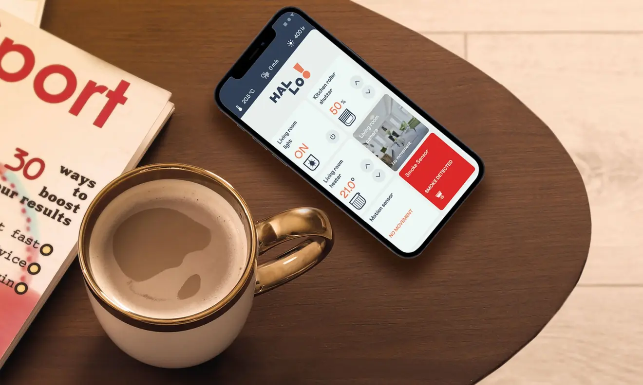 Smartphone auf Tisch neben Tasse und Zeitschrift, zeigt Smart-Home-App mit Steuerungsoptionen