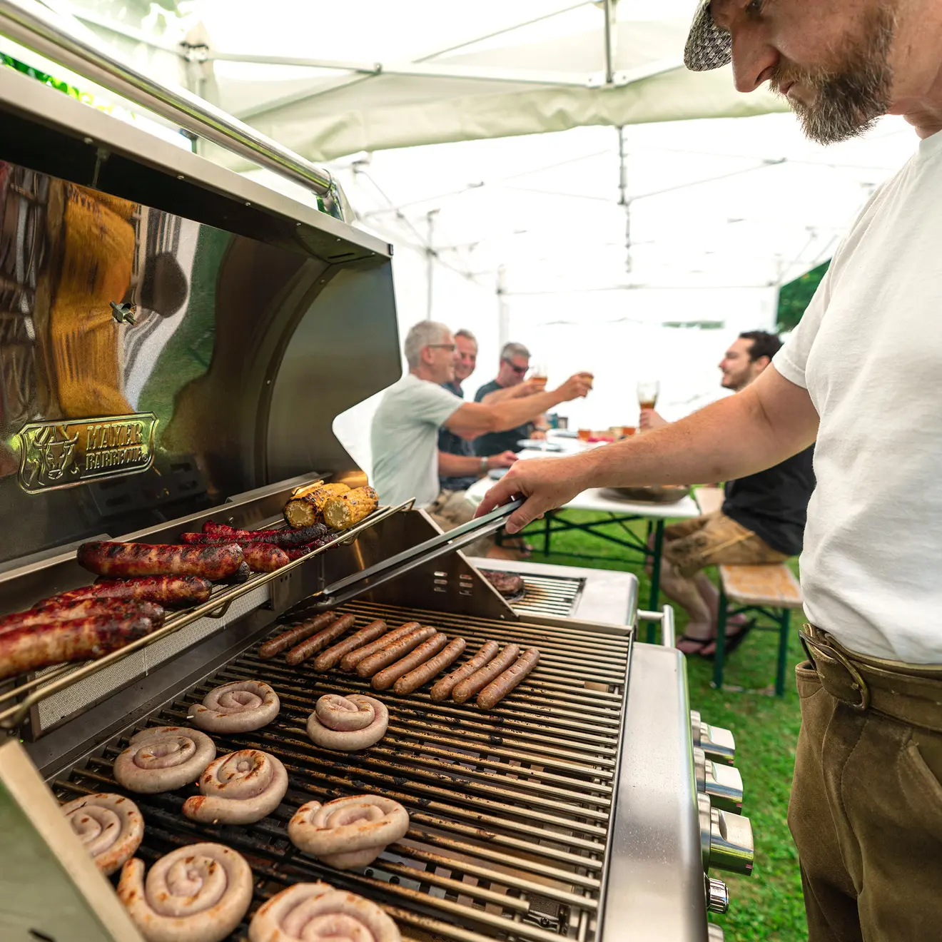 Detailbild von dem MGG-442 Mastergrill mit Fleisch auf dem Rost und Personen die im Hintergrund sitzen