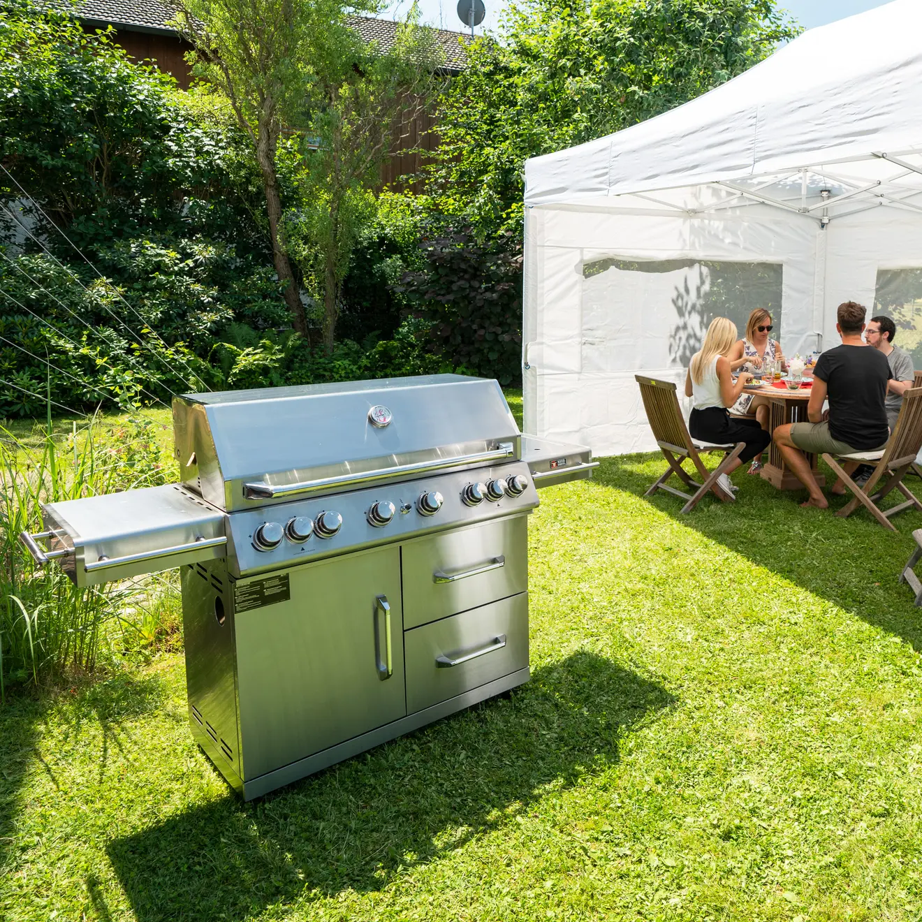 Grill im Garten, im Hintergrund Personen unter einem Pavillon beim essen