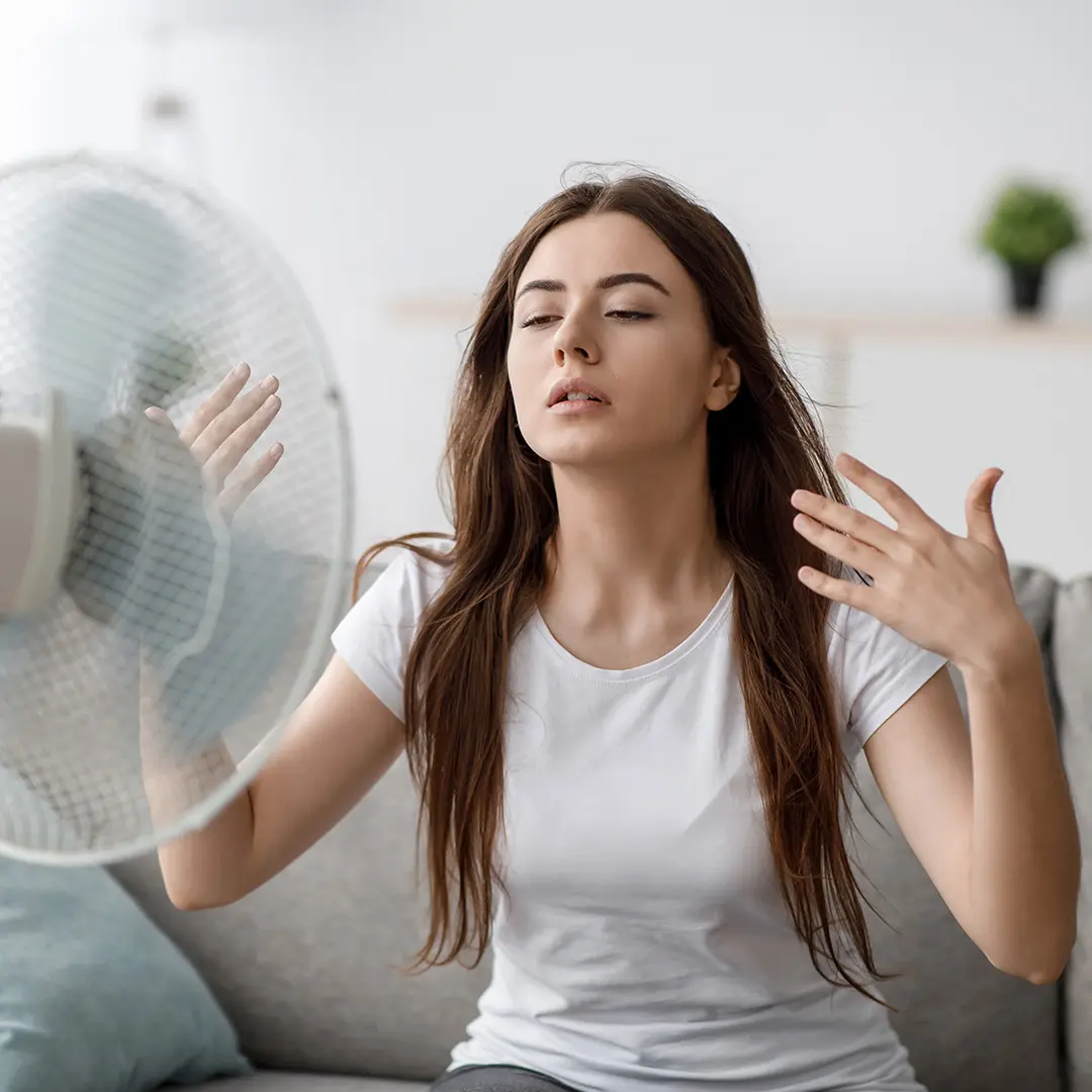 Junge Frau sitzt im Wohnzimmer vor einem Ventilator, der ihr kühlende Luft entgegenbläst.