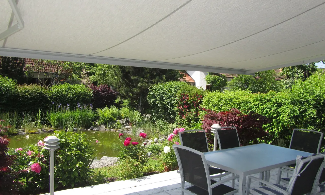 Gelenkarmmarkise Basic 2000 über Terrasse mit Tisch und Stühlen, Garten im Hintergrund