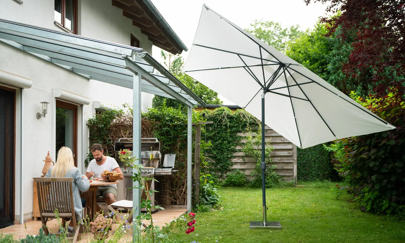 Terrasse mit großem Sonnenschirm und Personen am Tisch unter Glasüberdachung im Garten