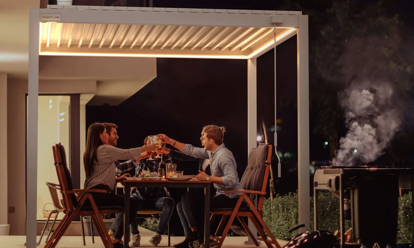 Menschen sitzen unter einer Pergola mit Lamellendach und stoßen an, Grill im Hintergrund