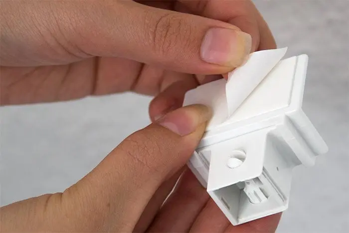Hand hält Klebehalter aus Befestigungs-Set für EasyFix Plissees, Schutzfolie wird abgezogen